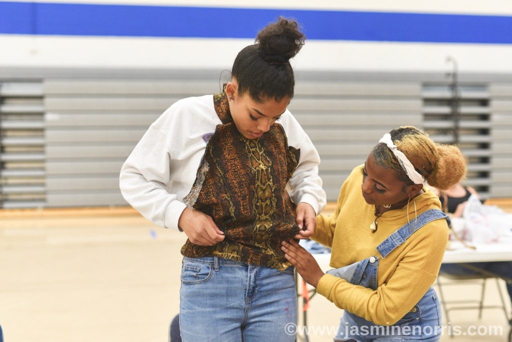 Two teenage girls, one wearing a yellow cropped sweater with light blue overalls draping a piece of fabric on the other teenage girl.